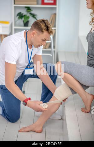 Fisioterapista maschile inginocchiato su un ginocchio, applicando bendaggio al ginocchio della paziente femminile Foto Stock