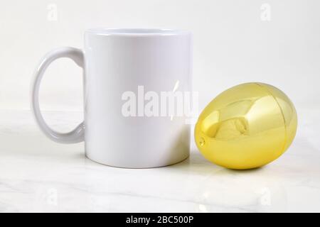 Questa tazza di caffè di Pasqua è caratterizzata da una tazza di caffè bianca da 11 g che si distacca su uno sfondo di marmo bianco. Accanto alla tazza a si trova un uovo di Pasqua d'oro Foto Stock