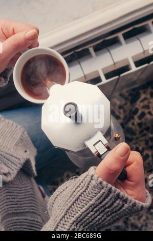 Donna che versa il caffè da una moka pentola per la colazione a casa di fronte a una finestra al mattino Foto Stock