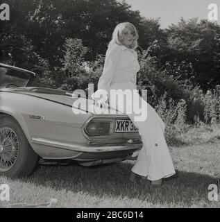1970s, storico, esterno in un campo illuminato dal sole, una donna bionda attraente in un top bianco e pantaloni a campana, in piedi accanto alla sua auto sportiva open-top, una Triumph Spitfire IV, con ruote a filo, Inghilterra, Regno Unito. Introdotto per la prima volta nel 1962, sulla base di un disegno di Giovanni Michelotti, lo Spitfire era una piccola vettura sportiva britannica a due posti e prodotta fino al 1980. Il modello Mark IV qui illustrato è stato realizzato presso le opere di Standard-Triumph (di proprietà di Leyland Motors) a Canley, Coventry tra il 1970 e il 1974 e, tra le altre novità dei modelli precedenti, è stato un'estremità posteriore di nuova progettazione Foto Stock