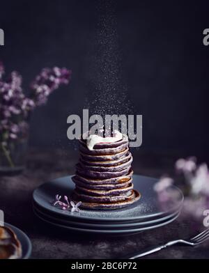 Frittelle viola con mirtilli e crema intorno fiori lilla a sfondo scuro atmosferico in primavera Foto Stock