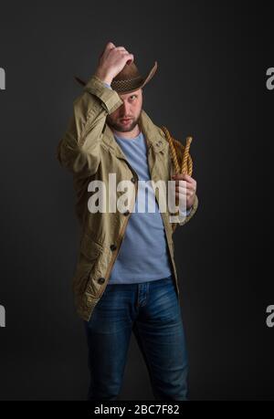 Cowboy in un cappello e una giacca a testa larga con un lazo sulla spalla che si posa su uno sfondo scuro Foto Stock