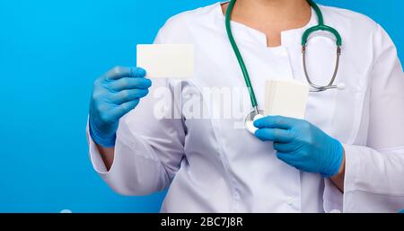 mano medico femmina in guanti di lattice medico blu tiene un biglietto da visita di carta vuoto su uno sfondo blu, luogo per un'iscrizione Foto Stock