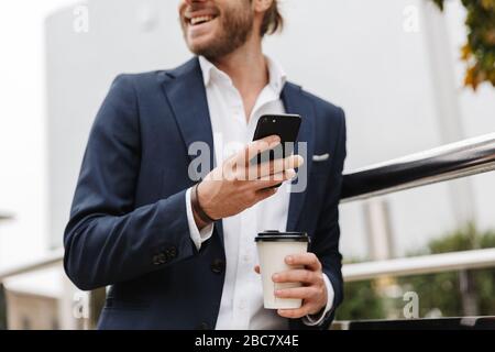 Immagine ritagliata di un giovane e sorridente uomo biondo dai capelli in formale indossare tenere takeaway caffè tazza e utilizzare il telefono cellulare mentre si cammina all'aperto Foto Stock