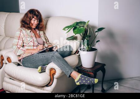 Felice donna di mezza età che lavora a casa su un tablet seduto sul divano in casa. Lavoro da casa concetto. Autoisolamento. Foto Stock