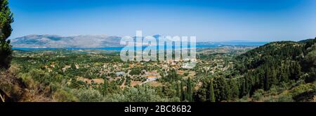 Vista panoramica ultra ampia dell'idilliaca cittadina a valle con tetti rossi sull'isola mediterranea. Oliveti, cipressi e baia blu in lontananza. Foto Stock