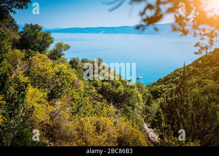 Vista panoramica ultra ampia dell'idilliaca cittadina a valle con tetti rossi sull'isola mediterranea. Oliveti, cipressi e baia blu in lontananza. Foto Stock