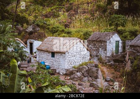 Case in pietra in stile locale con tetti coperti di paglia e finestre blu tra vegetazione lussureggiante e paesaggio montano. Santo Antao Capo Verde. Foto Stock