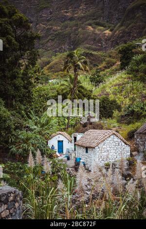 Case in pietra in stile locale con tetti coperti di paglia e finestre blu tra vegetazione lussureggiante e paesaggio montano. Santo Antao Capo Verde. Ve Foto Stock