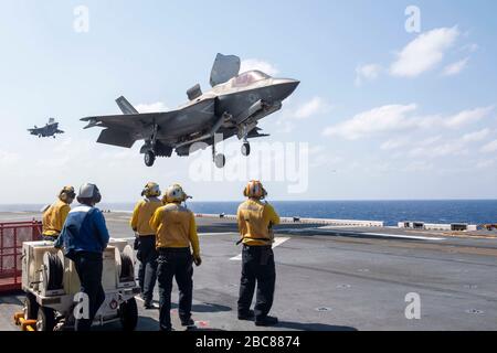 Una giacca gialla della marina statunitense guarda come un combattente furtivo contro i fulmini F-35B Lightning II, assegnato alla 31st Marine Expeditionary Unit, esegue un atterraggio verticale sul ponte di volo della nave d'assalto anfibio di Flagship America USS America durante le operazioni di routine del 25 marzo 2020 nel Mare delle Filippine. Foto Stock