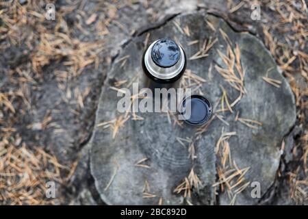 Bottiglia thermos nera e una tazza di tè in piedi su un moncone circondato da aghi caduti di pino, vista dall'alto. Picnic nel parco autunnale. Foto Stock