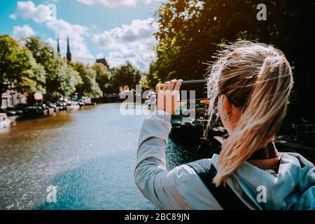 Femmina turistico scattare foto del canale di Amsterdam sul cellulare. Caldo oro pomeriggio luce del sole. Viaggi in Europa. Foto Stock