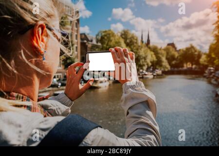 Donna turistica prendendo una foto del canale di Amsterdam sul telefono cellulare. Oro caldo pomeriggio di sole. Viaggiare in Europa. Foto Stock