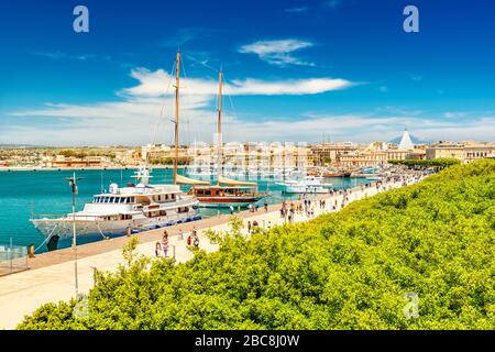 Splendida vista sul porto di Siracusa con persone che camminano lungo il lungomare. Paesaggio urbano dell'isola di Ortigia, Sicilia, Italia Foto Stock