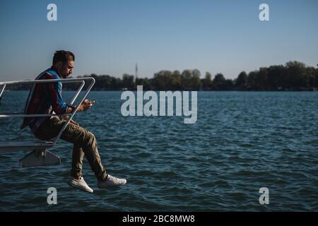 Serio bell'uomo che guarda il telefono cellulare mentre si siede sopra il fiume ona una bella giornata di sole Foto Stock
