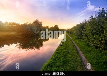 Guardando lungo un sentiero di stoppino canale inglese in una serata estiva, il sole tramonta con un bel cielo che si riflette nell'acqua Foto Stock