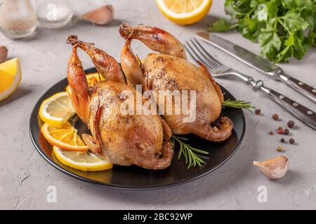 Quaglie al forno con limone e arancia servite su un piatto scuro su sfondo grigio Foto Stock