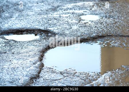 Vicino a una strada in pessime condizioni con grandi buche piene di piscine d'acqua piovana sporche. Foto Stock