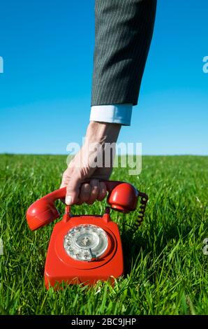 Mano di uomo d'affari irriconoscibile che solleva il ricevitore di un telefono a manopola rosso vecchio stile seduto all'aperto in erba verde Foto Stock