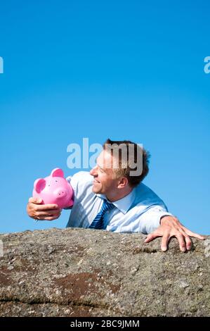 Imprenditore sorridente tenendo premuto su un fido rosa salvadanaio come egli raggiunge la cima di una scogliera rocciosa Foto Stock