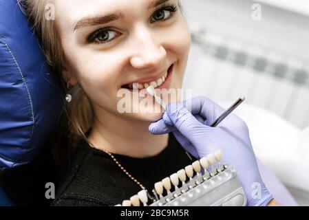 Controllo dentista del colore dei denti della paziente femmina, con scala di smalto dei denti, interno dell'ufficio dentale. La bionda sorride nella sedia dentale, in attesa della procedura di sbiancamento. Foto Stock