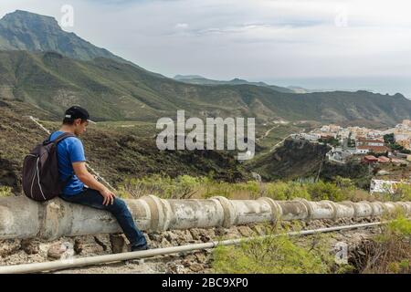 Un giovane con uno zaino si siedera sull'acquedotto in cemento nel lato ovest di Tenerife vicino al villaggio di Adeje. Escursioni lungo il sentiero di montagna circostante Foto Stock