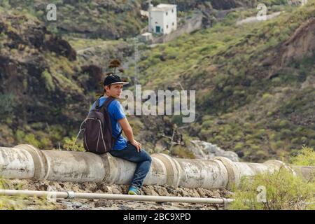 Un giovane con uno zaino si siedera sull'acquedotto in cemento nel lato ovest di Tenerife vicino al villaggio di Adeje. Escursioni lungo il sentiero di montagna circostante Foto Stock