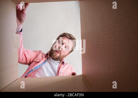 Sorpreso, l'uomo bearded soddisfatto scompatta una scatola consegnata con un pacco o un regalo. Disimballaggio della vista interna. Spazio di copia. Foto Stock