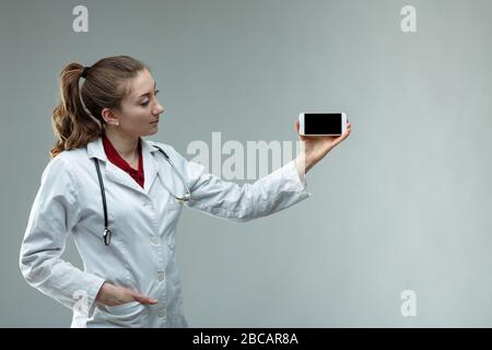 Donna medico o infermiere che indossa uno stetoscopio e un camice bianco da laboratorio in piedi che tiene il suo telefono cellulare con lo schermo alla fotocamera su copie grigie wth s. Foto Stock