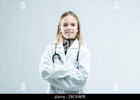 Sorridente ragazza amichevole vestita come un medico in un camice bianco da laboratorio con stetoscopio in piedi con le braccia piegate che grinning alla fotocamera su bianco con c. Foto Stock