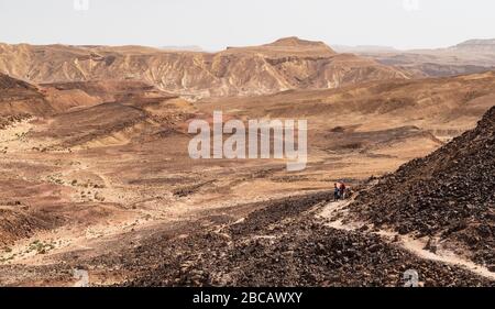 due escursionisti sulla collina di arut nel cratere makhtesh ramon in israele con rocce di arenaria ricoperte di patina in primo piano e il bordo del canyon sud Foto Stock