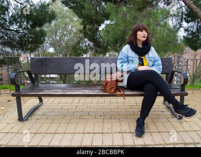 Ritratto di una giovane donna di stile urbano in abiti moderni e luminosi seduti su una panca. Stile di vita e concetto urbano. Foto Stock