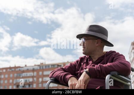 Uomo caucasico con occhiali e cappello depressivo e triste in una sedia a rotelle solitaria in un parco con il cielo e alcuni edifici sullo sfondo con lo spazio Foto Stock