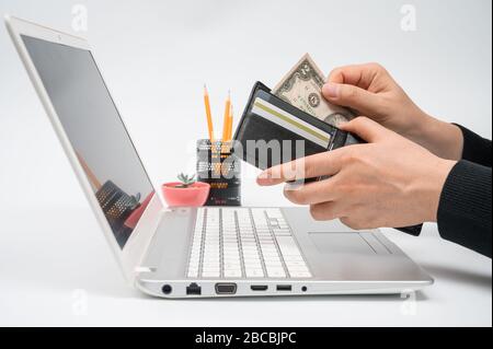 Concetto di e-commerce. La mano di un uomo estrae denaro dal portafoglio di fronte al monitor di un notebook. Foto Stock