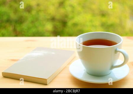 Tazza di tè caldo e un libro su un tavolo esterno con fogliame sfocato sullo sfondo Foto Stock