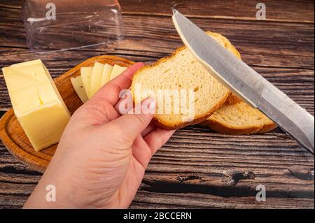 Qualcuno spalma il burro su pane tostato di grano fresco con un coltello, un piatto di burro di legno con un pezzo di burro e fette di formaggio su uno sfondo di legno. Clo Foto Stock