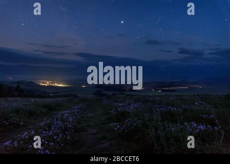 Paesaggio notturno con una strada sterrata in montagna, fiori di geranio in primo piano, luci del villaggio e tracce di fari da auto contro un bac Foto Stock