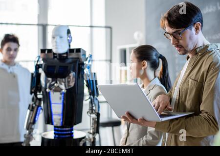 Giovane insegnante serio con laptop che guarda attraverso materiali online mentre i suoi studenti fanno la presentazione delle abilità robot al seminario Foto Stock