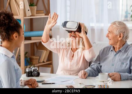 Agente di viaggio in ufficio con clienti senior donna che indossa cuffie per realtà virtuale godendo di un'esperienza allegra Foto Stock