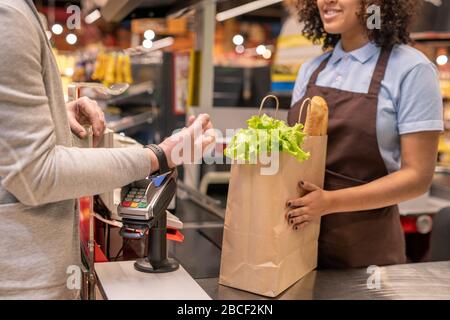 Mano di uomo maturo contemporaneo con smartwatch che tiene il polso sopra la macchina di pagamento mentre in piedi dal registratore di cassa nel supermercato Foto Stock