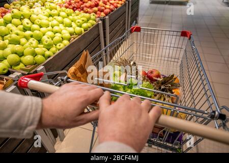 Mani di cliente maschio invecchiato che spingono il carrello con i prodotti alimentari mentre si spostano lungo l'esposizione della frutta con le mele fresche nel supermercato Foto Stock