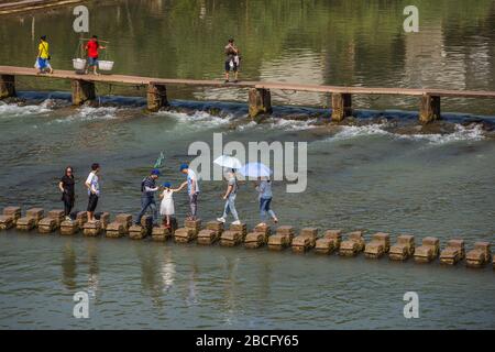 Feng Huang, Cina - Agosto 2019 : persone che attraversano l'acqua su pietre a passo sul fiume Tuo Tuojiang, che scorre attraverso il centro della città vecchia Fenghuang, H. Foto Stock