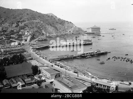 Vista ad alto angolo di Avalon Bay, Avalon, Santa Catalina Island, California, 1931. Nel porto sono ormeggiate barche e una nave a vapore attraccata presso un molo, le persone si costeggiano le spiagge e si sfilano in acqua, e la strada lungomare conduce al Catalina Casino Ballroom. Fotografia di Burton Holmes. Foto Stock