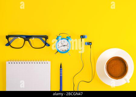 Piccola sveglia blu, in stile minimalista, auricolari blu, occhiali blu e libro di note con penna blu, si trova su sfondo giallo brillante. Caffè cu Foto Stock