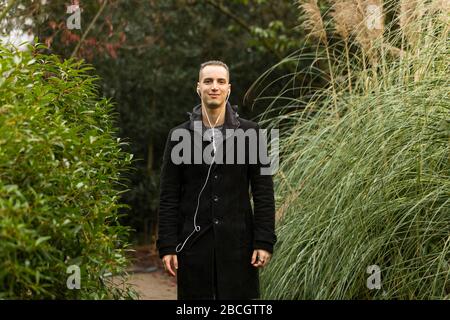 Giovane uomo in posa in giardino indossando cappotto e ascoltando le cuffie Foto Stock
