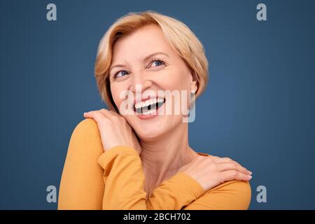 Stile libero. Donna matura in piedi isolata su blu abbracciandosi ridendo gioiosa primo piano Foto Stock