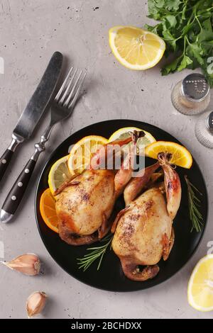 Quaglie al forno con limone e arancio servite su un piatto scuro su sfondo grigio, vista dall'alto, orientamento verticale Foto Stock