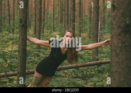 Ritratto di giovane bella ragazza in abito nero in foresta che raggiunge tronco di albero Foto Stock