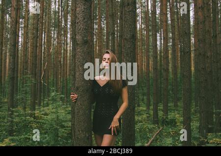 Ritratto di giovane bella ragazza in abito nero in foresta che tiene tronco di albero Foto Stock