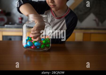 il bambino prende i cioccolatini colorati dal vaso trasparente in cucina Foto Stock